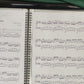 Close-up video of a girl using a piano display folder as she removes a pencil from the attached holder, carefully adds a sharp symbol next to a note on the sheet music, and then places the pencil back in the holder. The music folder keeps the sheet steady and organized for easy marking